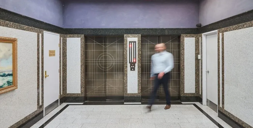 lift-background-mobile Art deco elevator lobby with two metal elevator doors and a blurred person walking across the tiled floor.
